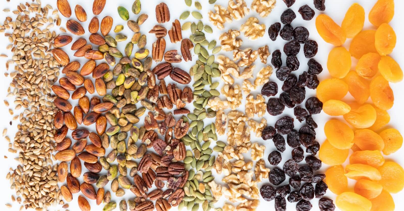 A top-view photo displays an assortment of nuts, seeds, and dried fruits arranged in vertical rows. From left to right: sunflower seeds, almonds, pistachios, pecans, pumpkin seeds, walnuts, dried cranberries, and dried apricots—all sourced from top nut suppliers in Metro Manila—set against a white background.