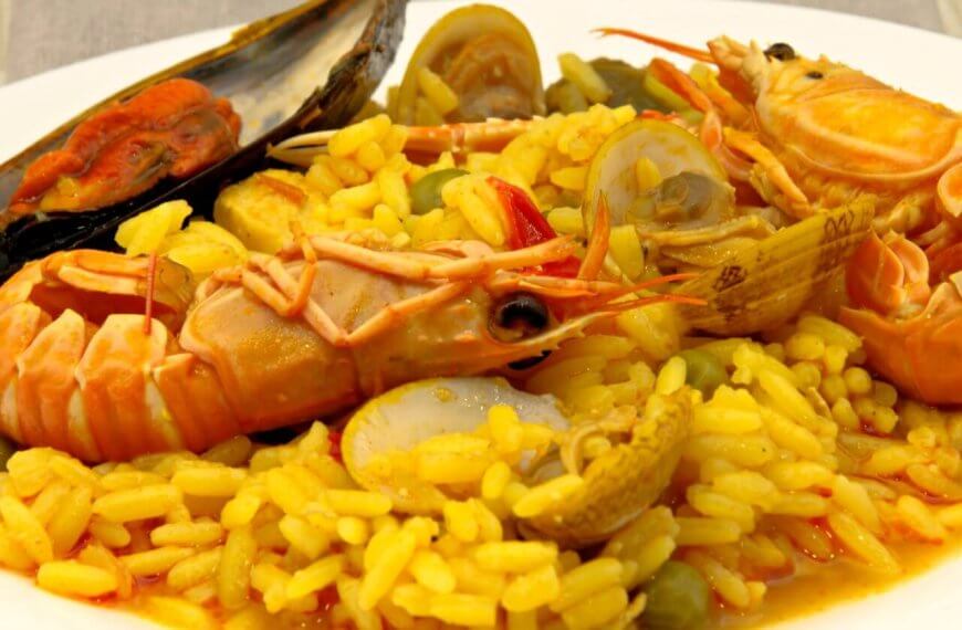 A plate of vibrant yellow paella, garnished with large prawns, clams, a mussel, and green peas. The rice is infused with saffron, giving it a rich color and flavor, and is interspersed with small chunks of vegetables. This top-notch dish showcases an assortment of fresh seafood from one of Estancia Mall's finest restaurants.