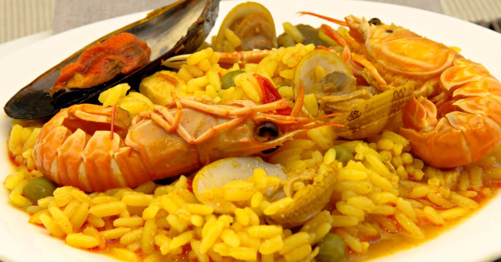A plate of vibrant yellow paella, garnished with large prawns, clams, a mussel, and green peas. The rice is infused with saffron, giving it a rich color and flavor, and is interspersed with small chunks of vegetables. This top-notch dish showcases an assortment of fresh seafood from one of Estancia Mall's finest restaurants.