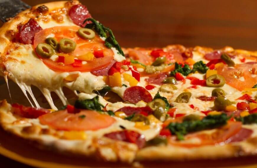 pizza at one of the best restaurants in Festival Mall. The pizza is topped with melted cheese, pepperoni, tomato slices, green olives, red bell peppers, and leafy greens. The crust is golden brown, and the cheese is stretchy as the slice is pulled away.