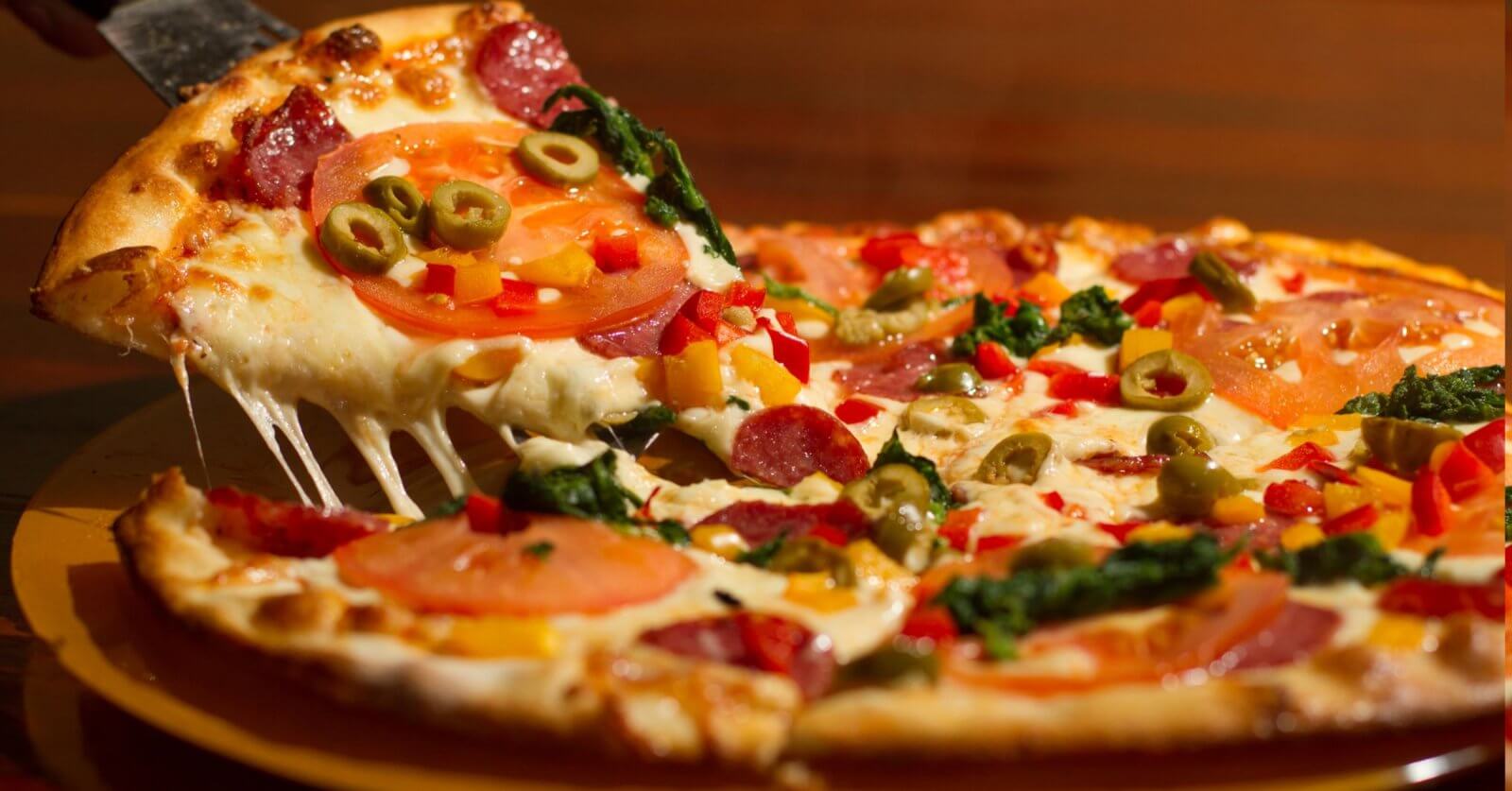 pizza at one of the best restaurants in Festival Mall. The pizza is topped with melted cheese, pepperoni, tomato slices, green olives, red bell peppers, and leafy greens. The crust is golden brown, and the cheese is stretchy as the slice is pulled away.