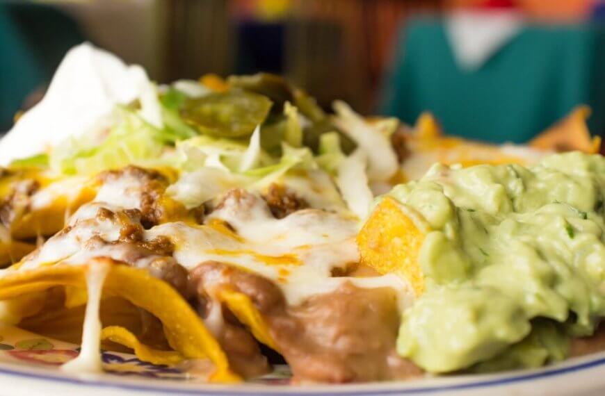 nachos topped with melted cheese, refried beans, ground beef, shredded lettuce, jalapeños, and a dollop of sour cream. On the side is a generous portion of creamy guacamole. The background is blurred to highlight the focus on these FIL-MEX delights from Metro Manila.