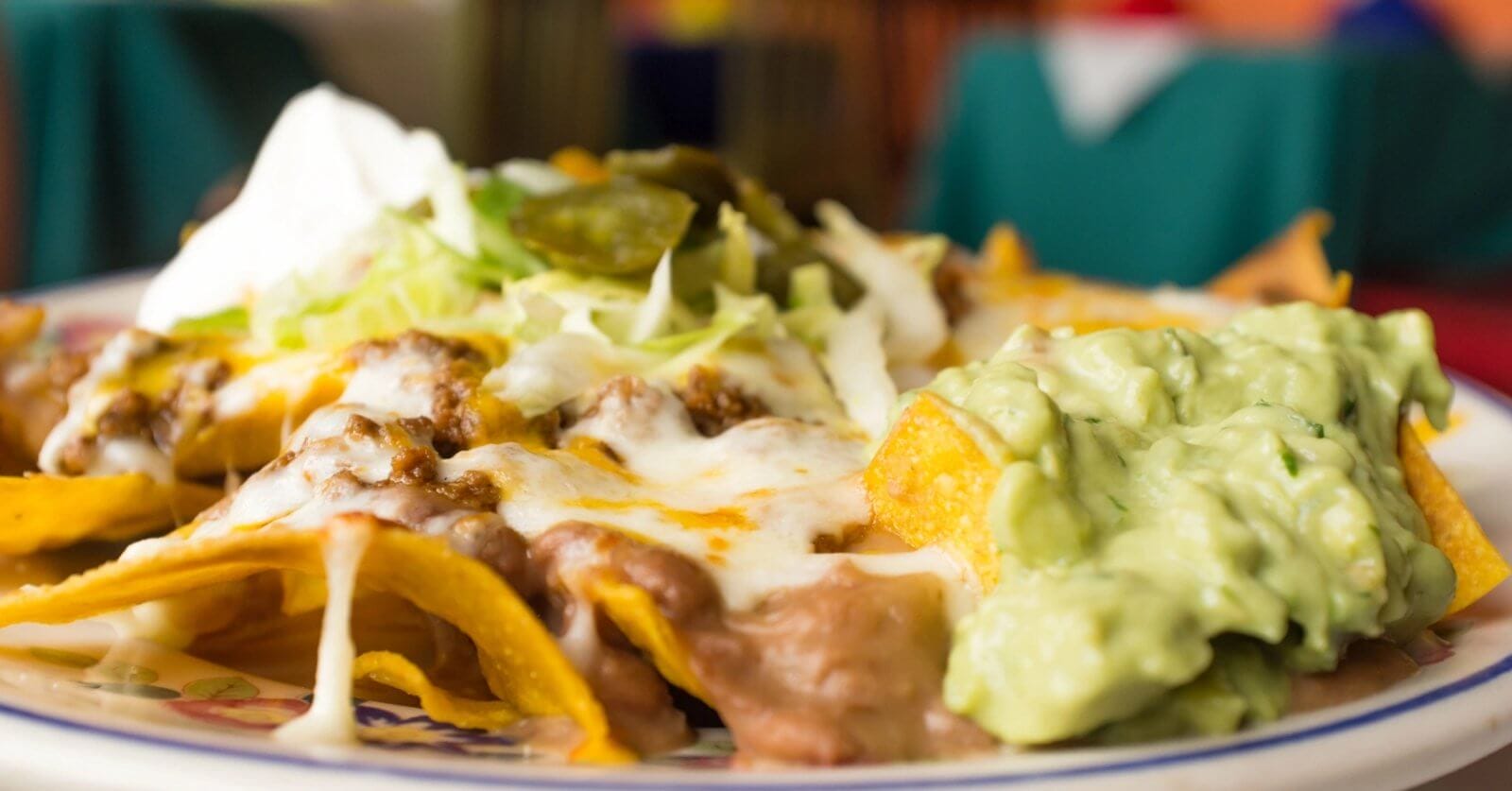 nachos topped with melted cheese, refried beans, ground beef, shredded lettuce, jalapeños, and a dollop of sour cream. On the side is a generous portion of creamy guacamole. The background is blurred to highlight the focus on these FIL-MEX delights from Metro Manila.