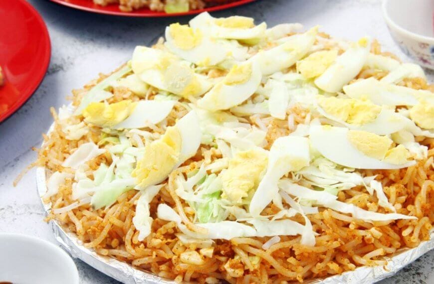 A plate of Filipino pancit palabok is topped with sliced hard-boiled eggs, shredded cabbage, and bits of garlic. The yellow-orange noodles are seasoned and mixed with shrimp sauce. Nearby, there are partially visible plates and bowls with other dishes and condiments on a white textured surface, reminiscent of the vibrant food parks in Metro Manila.