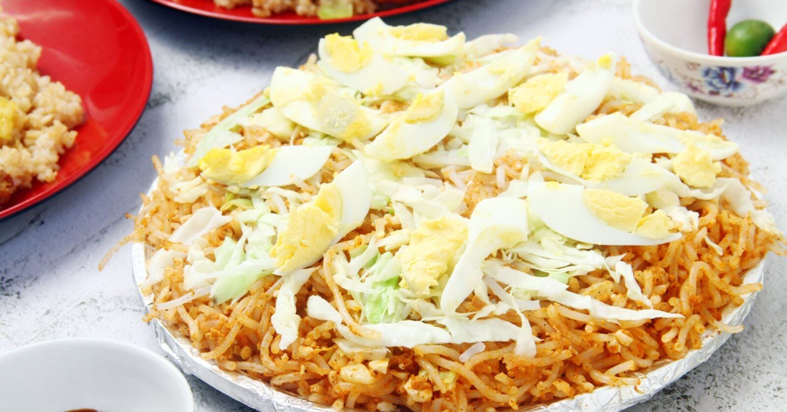 A plate of Filipino pancit palabok is topped with sliced hard-boiled eggs, shredded cabbage, and bits of garlic. The yellow-orange noodles are seasoned and mixed with shrimp sauce. Nearby, there are partially visible plates and bowls with other dishes and condiments on a white textured surface, reminiscent of the vibrant food parks in Metro Manila.