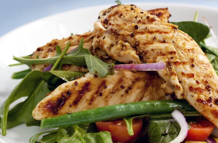 A plate of grilled chicken breasts served on a bed of fresh mixed greens, including arugula and green beans, is now available through healthy food delivery in Manila. The dish is garnished with slices of red onion and halves of cherry tomatoes. The chicken has a seared, seasoned exterior, and the salad appears crisp and vibrant.
