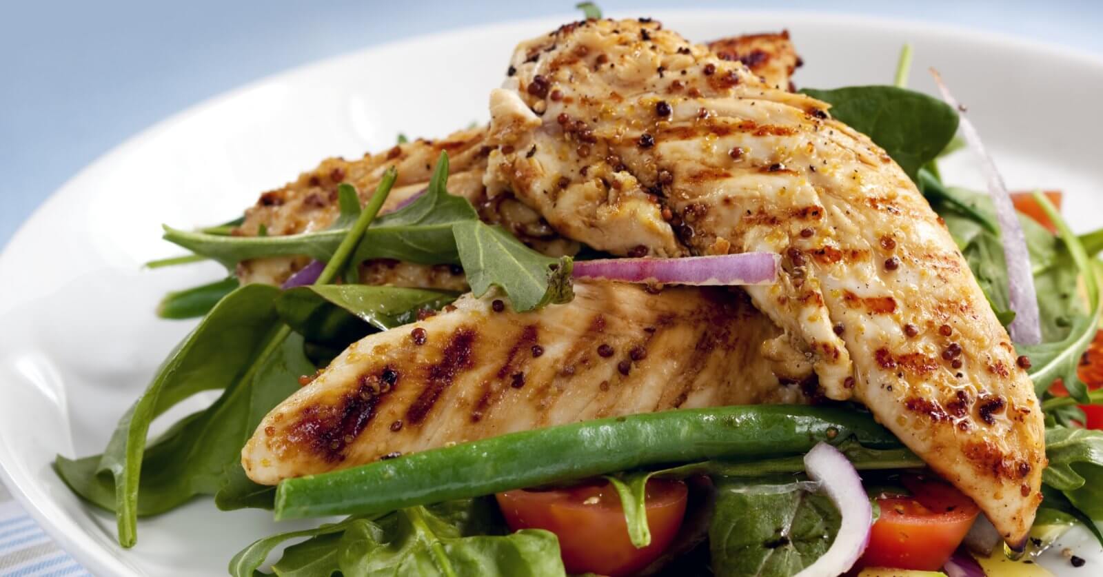 A plate of grilled chicken breasts served on a bed of fresh mixed greens, including arugula and green beans, is now available through healthy food delivery in Manila. The dish is garnished with slices of red onion and halves of cherry tomatoes. The chicken has a seared, seasoned exterior, and the salad appears crisp and vibrant.