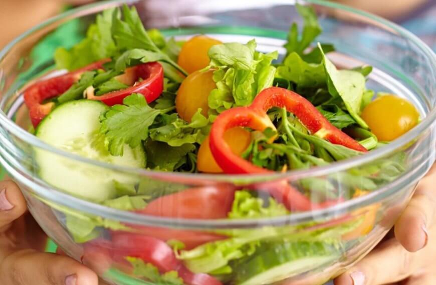A person is holding a clear glass bowl filled with a fresh salad. The salad includes slices of red bell pepper, rounds of cucumber, yellow cherry tomatoes, arugula, and mixed greens. Another person wearing a blue and green striped shirt is partially visible in the background at one of Metro Manila's healthy restaurants.