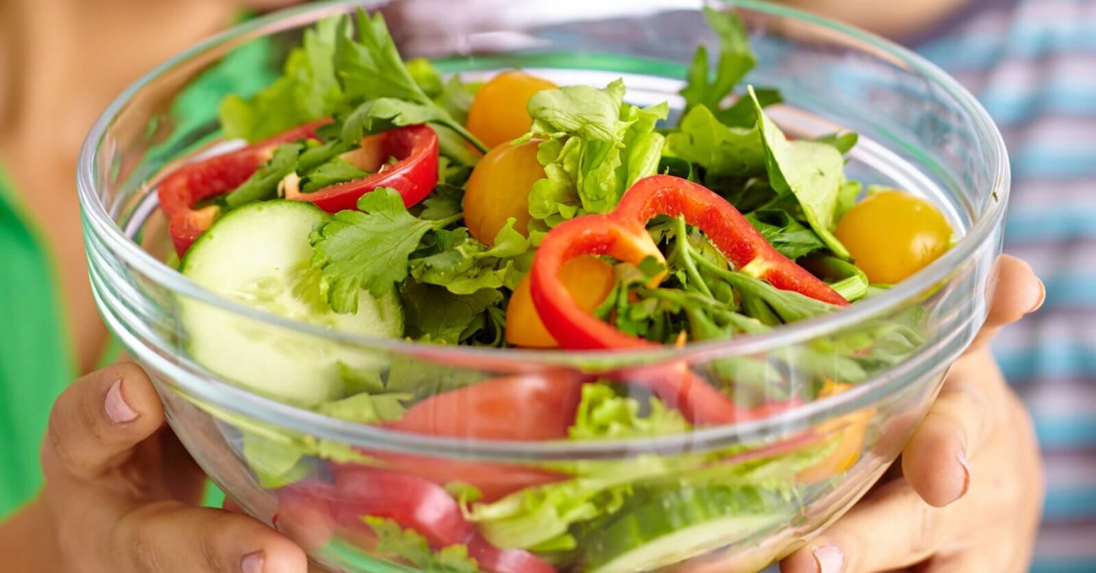 A person is holding a clear glass bowl filled with a fresh salad. The salad includes slices of red bell pepper, rounds of cucumber, yellow cherry tomatoes, arugula, and mixed greens. Another person wearing a blue and green striped shirt is partially visible in the background at one of Metro Manila's healthy restaurants.