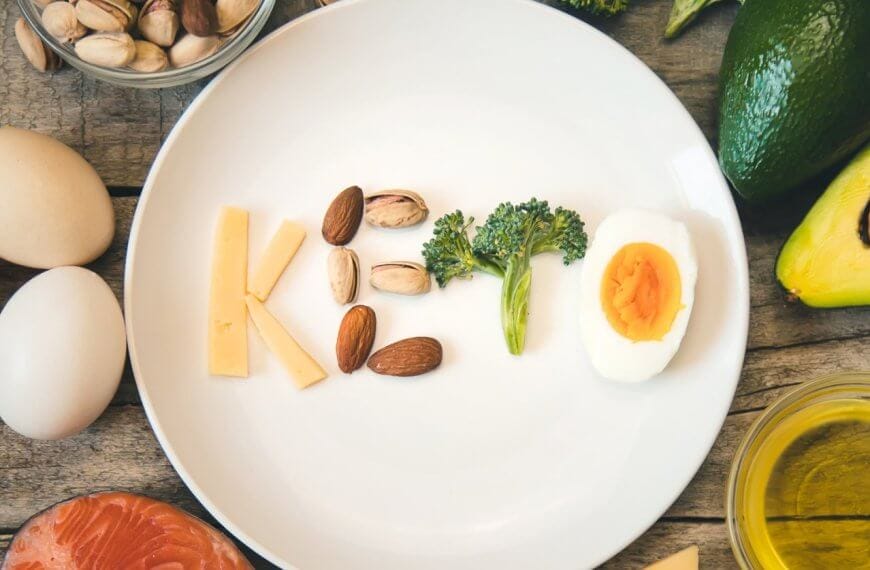 A white plate on a wooden table spells out "Keto" using various foods: cheese for "K", almonds and pistachios for "e", broccoli for "t", and a deviled egg for "o". Surrounding the plate are keto-friendly ingredients like eggs, an avocado, salmon, nuts, broccoli, and a bowl of oil. Discover fresh keto supplies in Metro Manila shops.