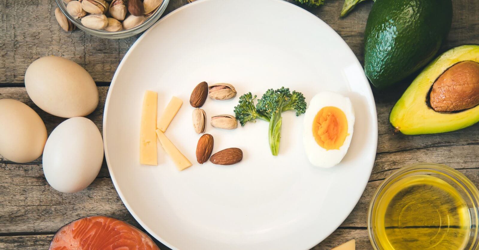A white plate on a wooden table spells out "Keto" using various foods: cheese for "K", almonds and pistachios for "e", broccoli for "t", and a deviled egg for "o". Surrounding the plate are keto-friendly ingredients like eggs, an avocado, salmon, nuts, broccoli, and a bowl of oil. Discover fresh keto supplies in Metro Manila shops.