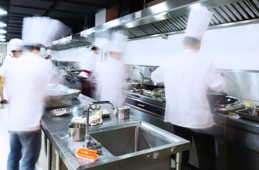 A bustling commercial kitchen with stainless steel counters, sinks, and ventilation hoods. Several chefs in white uniforms and hats are blurred, indicating rapid movement as they address every culinary need. Pots, pans, and utensils from Top 50 Kitchen Supply Stores in Metro Manila are scattered across the workspaces.
