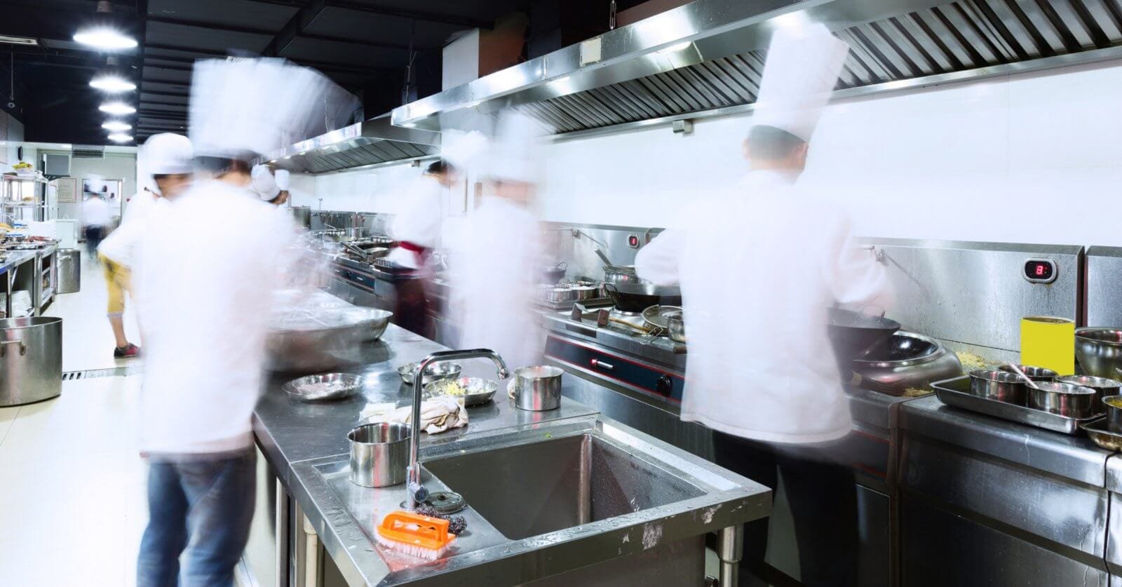 A bustling commercial kitchen with stainless steel counters, sinks, and ventilation hoods. Several chefs in white uniforms and hats are blurred, indicating rapid movement as they address every culinary need. Pots, pans, and utensils from Top 50 Kitchen Supply Stores in Metro Manila are scattered across the workspaces.