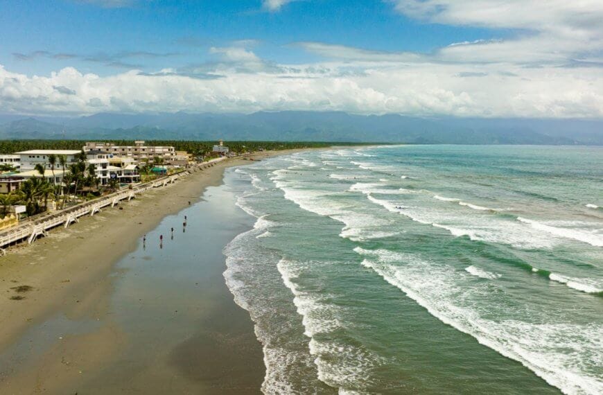 An aerial view of a long, sandy beach on Luzon with gentle waves rolling in from a turquoise ocean. On the left side, buildings and palm trees that are popular tourist attractions line the shore, while a few people are scattered along the wet sand. In the background, lush mountains are partially obscured by clouds under a bright, blue sky.
