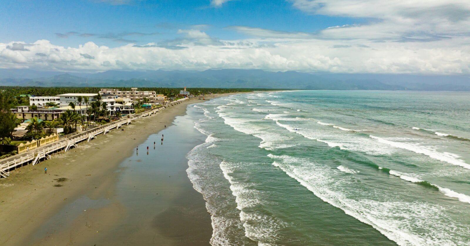 An aerial view of a long, sandy beach on Luzon with gentle waves rolling in from a turquoise ocean. On the left side, buildings and palm trees that are popular tourist attractions line the shore, while a few people are scattered along the wet sand. In the background, lush mountains are partially obscured by clouds under a bright, blue sky.