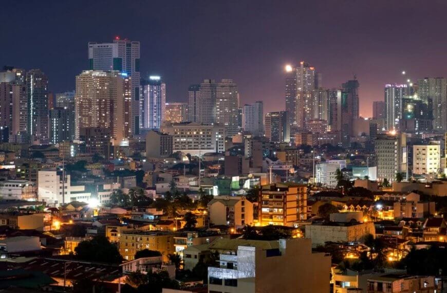 Makati cityscape at dusk featuring numerous high-rise buildings illuminated by varied lights defines the bustling spirit of Makati. The foreground consists of lower residential buildings with streetlights glowing, near Barangay Halls. The sky transitions from purple to blue, reflecting off some skyscrapers.