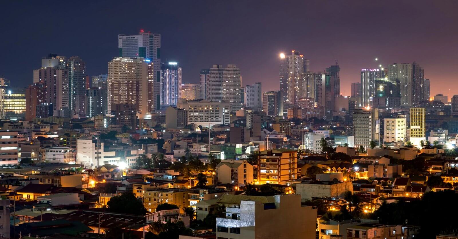 Makati cityscape at dusk featuring numerous high-rise buildings illuminated by varied lights defines the bustling spirit of Makati. The foreground consists of lower residential buildings with streetlights glowing, near Barangay Halls. The sky transitions from purple to blue, reflecting off some skyscrapers.