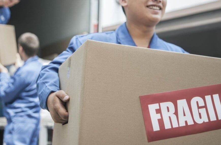 A person smiling and holding a large cardboard box with a red "FRAGILE" label. In the background, another person in a similar uniform is unloading a box from a truck. The setting appears to be one of the best moving companies in Metro Manila, providing top-notch delivery service.