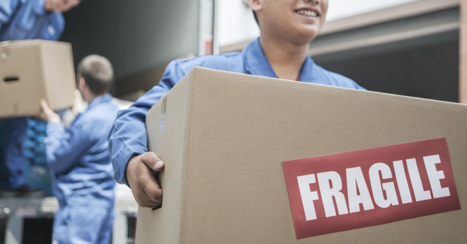 A person smiling and holding a large cardboard box with a red "FRAGILE" label. In the background, another person in a similar uniform is unloading a box from a truck. The setting appears to be one of the best moving companies in Metro Manila, providing top-notch delivery service.