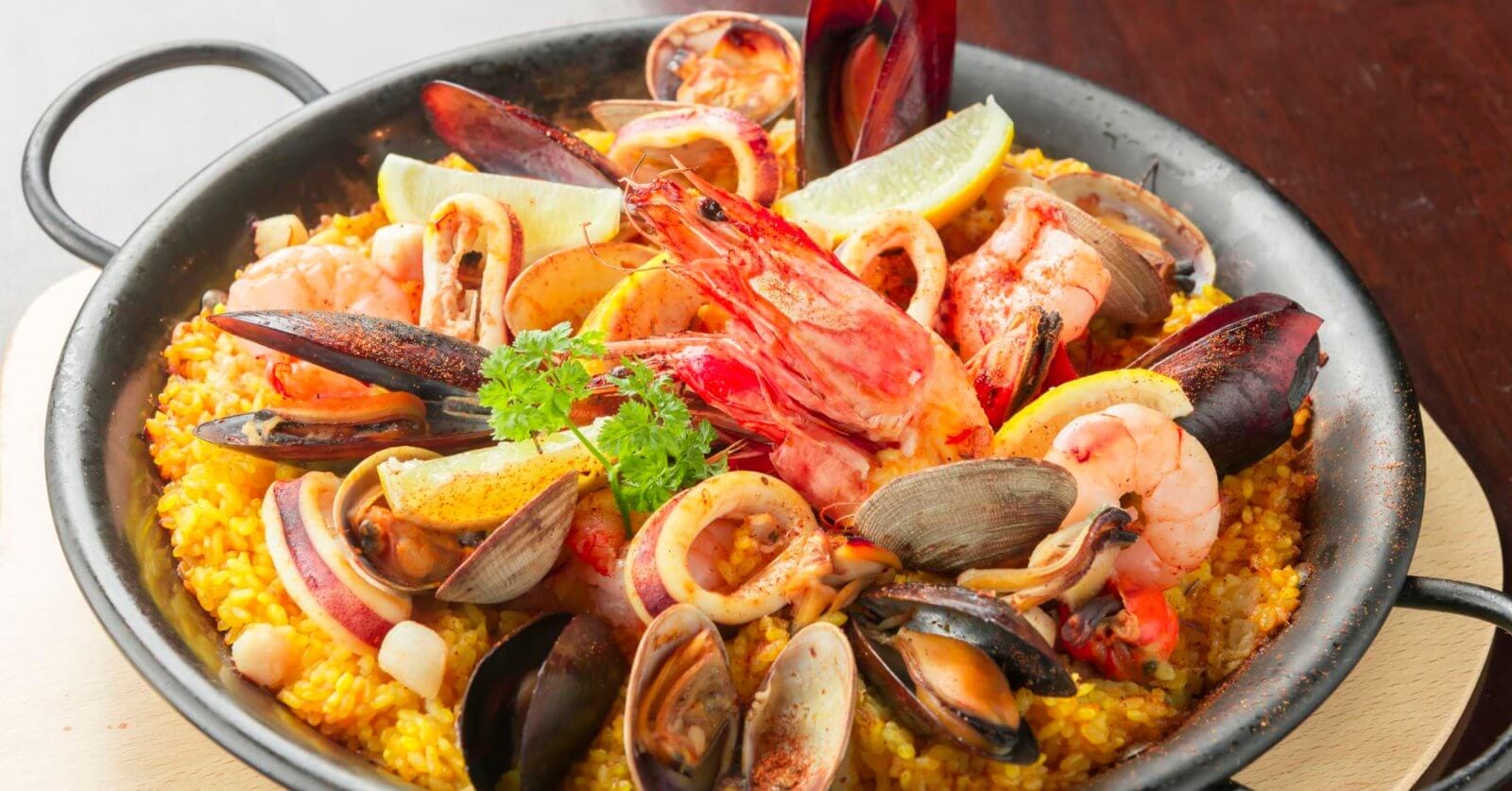 A black skillet filled with vibrant Spanish seafood paella showcases a sulit dining experience. The dish features yellow saffron rice topped generously with mussels, clams, shrimp, squid, and slices of lemon. A sprig of parsley garnishes the center. Ideal for those exploring the best restaurants in Muntinlupa City.