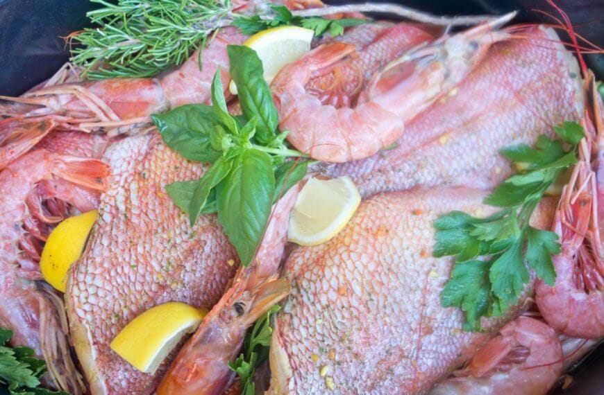 A seafood dish featuring whole fish and shrimp garnished with fresh herbs, including parsley and rosemary, is displayed. Lemon wedges are scattered throughout the arrangement, adding a vibrant contrast to the pink and red hues of the seafood, reminiscent of a Panglao feast.