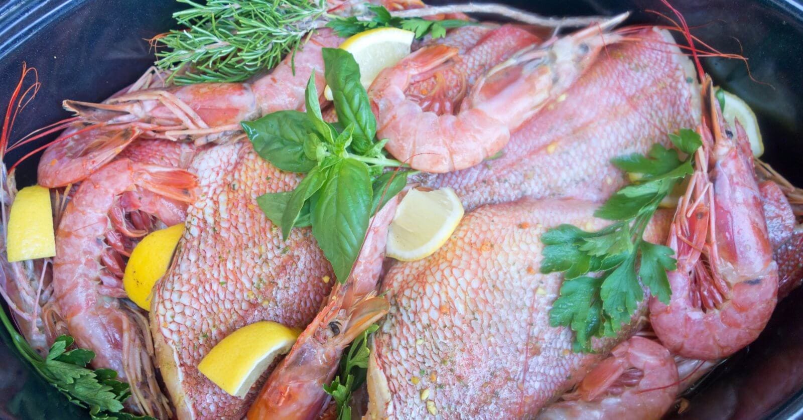 A seafood dish featuring whole fish and shrimp garnished with fresh herbs, including parsley and rosemary, is displayed. Lemon wedges are scattered throughout the arrangement, adding a vibrant contrast to the pink and red hues of the seafood, reminiscent of a Panglao feast.