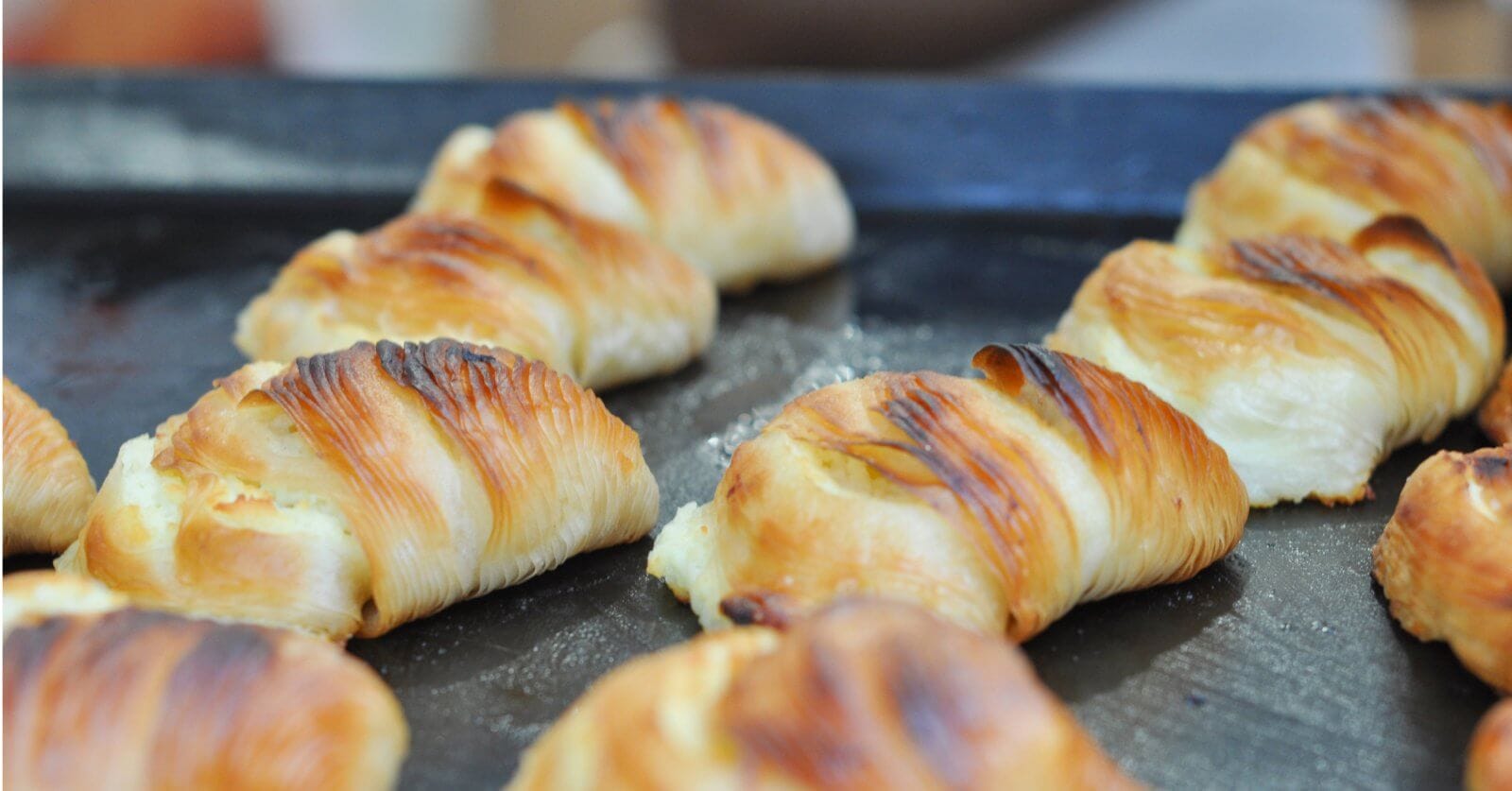 A close-up of golden-brown croissants lined up on a dark baking tray. The croissants appear freshly baked with flaky, buttery layers and slightly crispy, browned tops. Perfect for those sweet cravings, these treats are sourced from top pastry suppliers in Metro Manila.