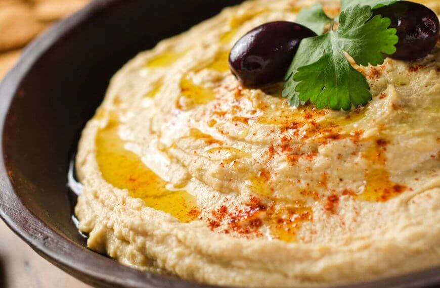 A close-up shot of a bowl of creamy hummus, garnished with two black olives, a sprig of fresh parsley, and a drizzle of olive oil. A sprinkle of paprika adds a touch of color on top. The dish is presented in a dark bowl, with blurred crackers visible in the background—a true culinary adventure.