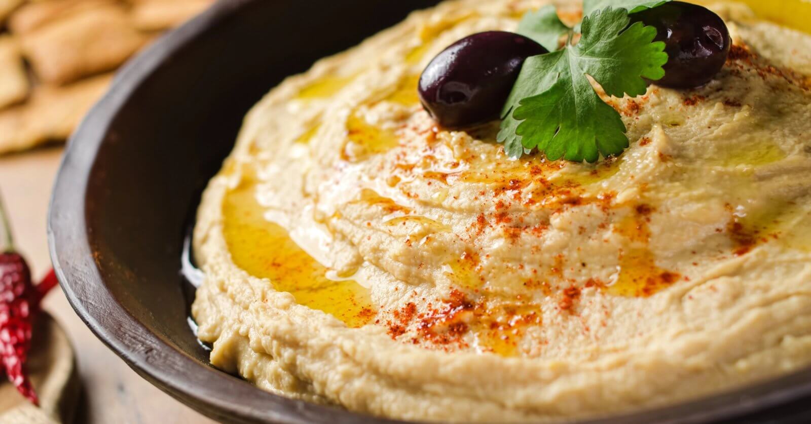 A close-up shot of a bowl of creamy hummus, garnished with two black olives, a sprig of fresh parsley, and a drizzle of olive oil. A sprinkle of paprika adds a touch of color on top. The dish is presented in a dark bowl, with blurred crackers visible in the background—a true culinary adventure.