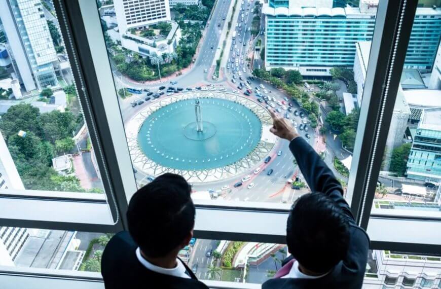 Two individuals in business attire are standing beside large windows in a high-rise building, overlooking the bustling cityscape of Metro Manila. One person is pointing toward a circular fountain surrounded by traffic on a busy street below. Tall buildings, greenery, and various vehicles are visible, highlighting the prime locations often sought by realty companies.