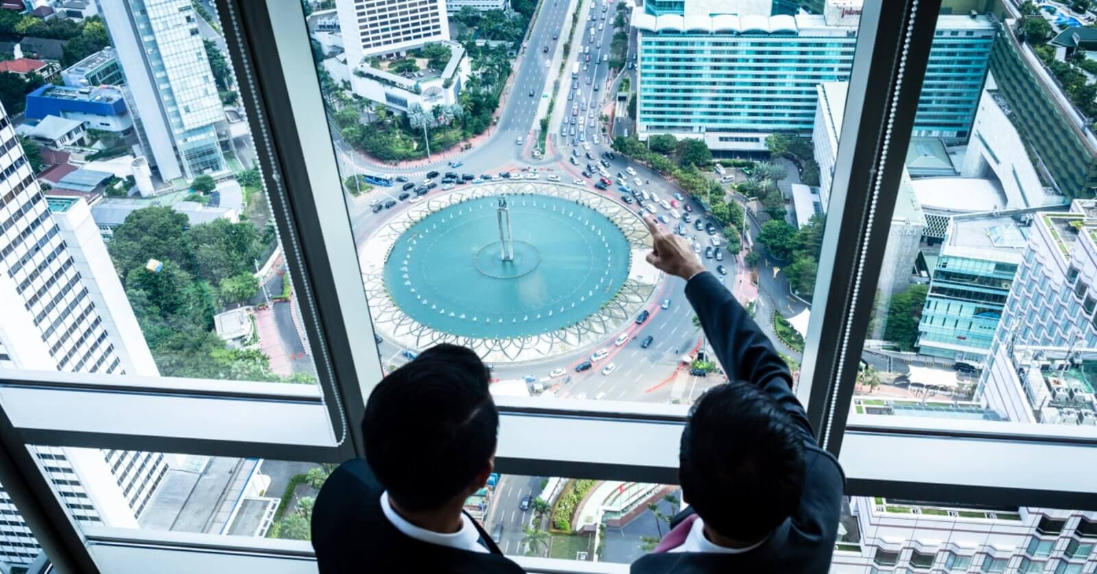 Two individuals in business attire are standing beside large windows in a high-rise building, overlooking the bustling cityscape of Metro Manila. One person is pointing toward a circular fountain surrounded by traffic on a busy street below. Tall buildings, greenery, and various vehicles are visible, highlighting the prime locations often sought by realty companies.