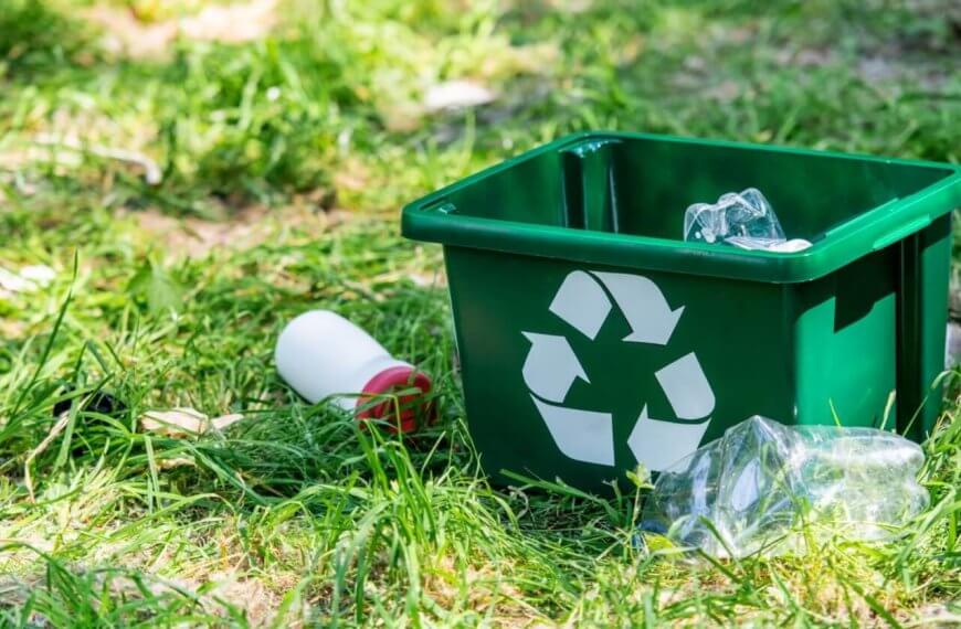 A green recycling bin with a white recycling symbol lies on the grass outdoors in Metro Manila. The bin contains a crumpled plastic bottle and is surrounded by scattered litter, including another crumpled plastic bottle and a white bottle with a red cap. Sunlight filters through the grassy area.