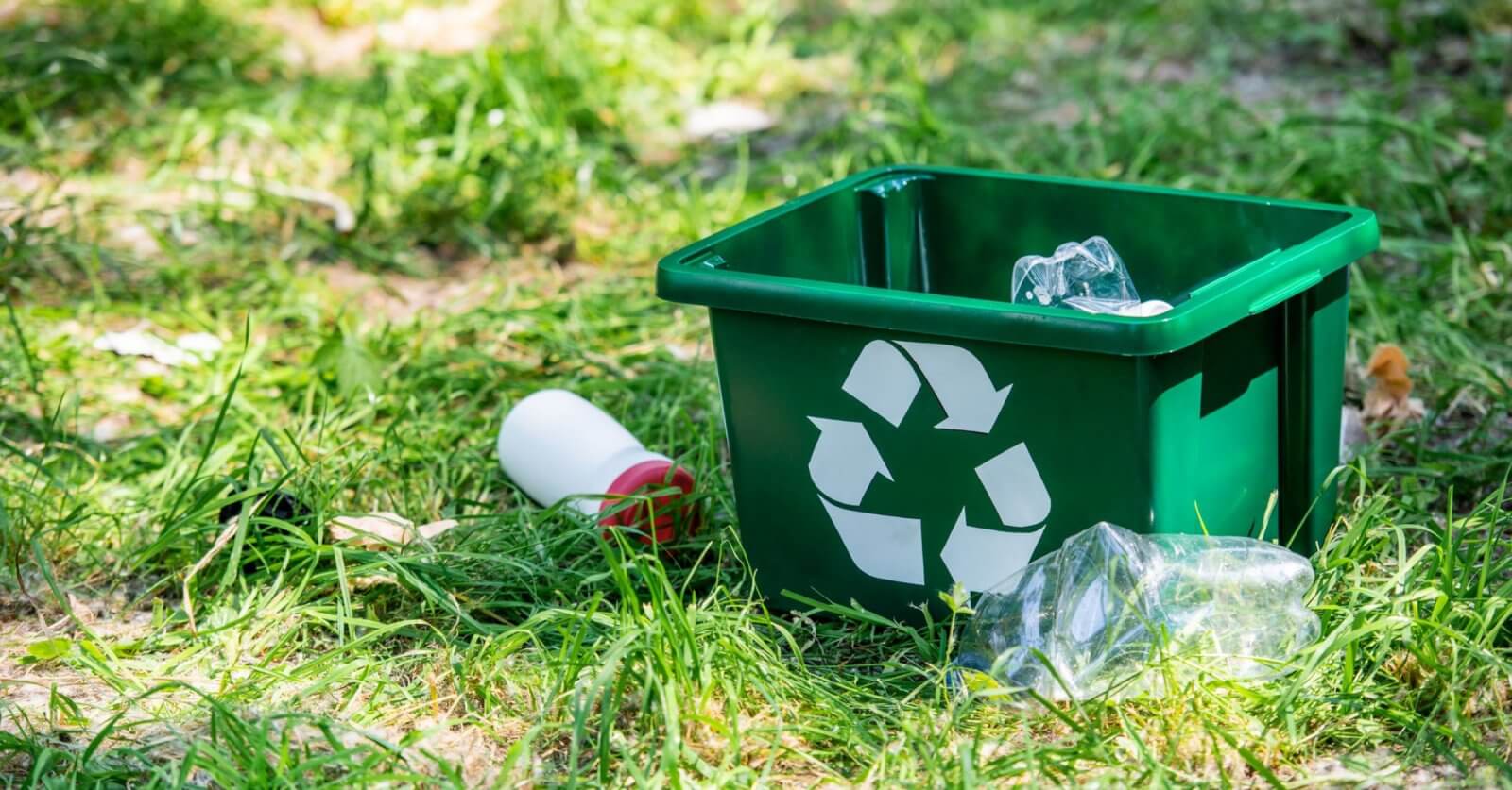 A green recycling bin with a white recycling symbol lies on the grass outdoors in Metro Manila. The bin contains a crumpled plastic bottle and is surrounded by scattered litter, including another crumpled plastic bottle and a white bottle with a red cap. Sunlight filters through the grassy area.