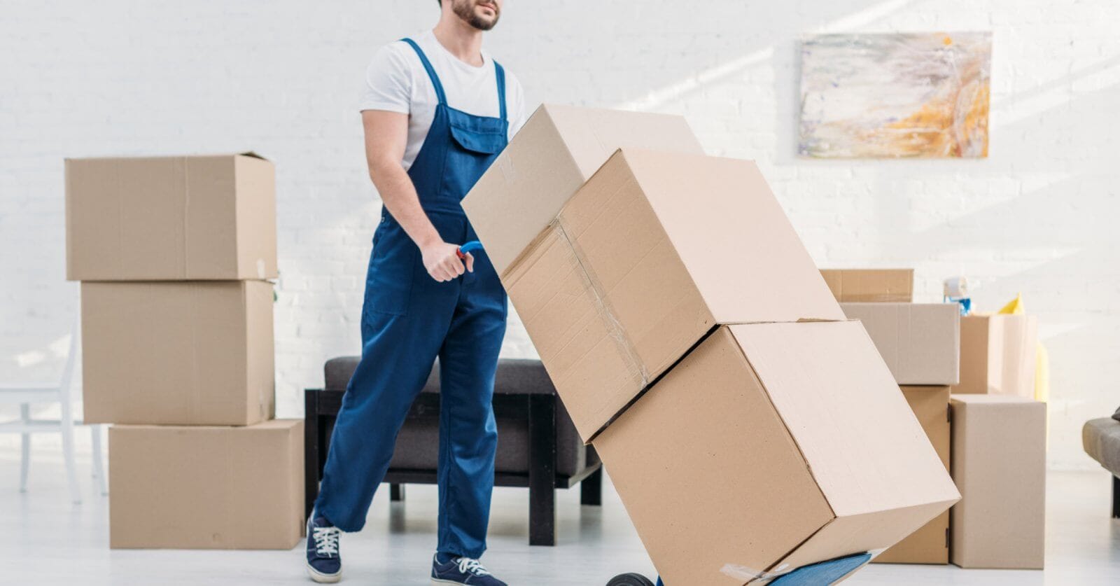 A person wearing a white t-shirt and blue overalls pushes a hand truck loaded with three large cardboard boxes. Several more boxes are stacked in the background of the room decorated with white walls and a modern abstract painting, indicating the provided relocation services. The person appears to be engaged in moving or packing activities.