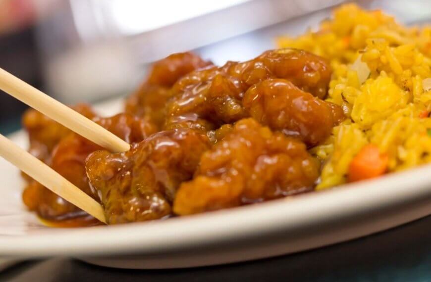 A plate featuring a serving of sweet and sour chicken, with pieces glistening in sauce and being picked up with a pair of chopsticks. On the side is a portion of fried rice, colorful with bits of vegetables like carrots and peas. This dish could be found at one of the best restaurants near Manila's 168 Shopping Mall.