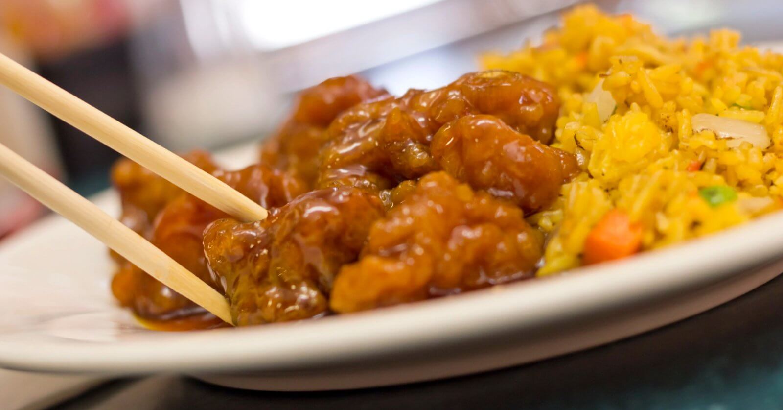 A plate featuring a serving of sweet and sour chicken, with pieces glistening in sauce and being picked up with a pair of chopsticks. On the side is a portion of fried rice, colorful with bits of vegetables like carrots and peas. This dish could be found at one of the best restaurants near Manila's 168 Shopping Mall.