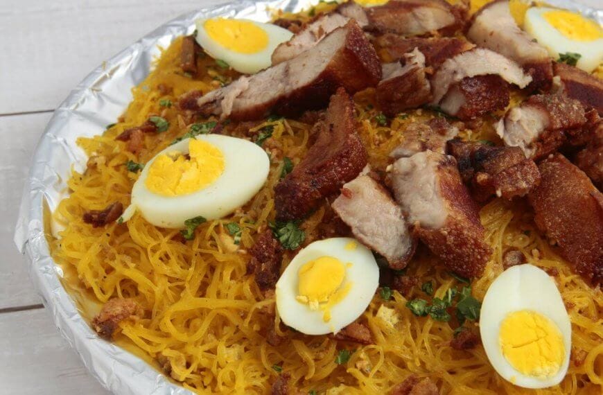 A plate filled with a Filipino noodle dish called Pancit Malabon, featuring yellow rice noodles topped with sliced hard-boiled eggs, crispy pork belly, cooked shrimp, and chopped green onions. The elements are arranged aesthetically on a white wooden table near the popular restos along Taft Avenue.