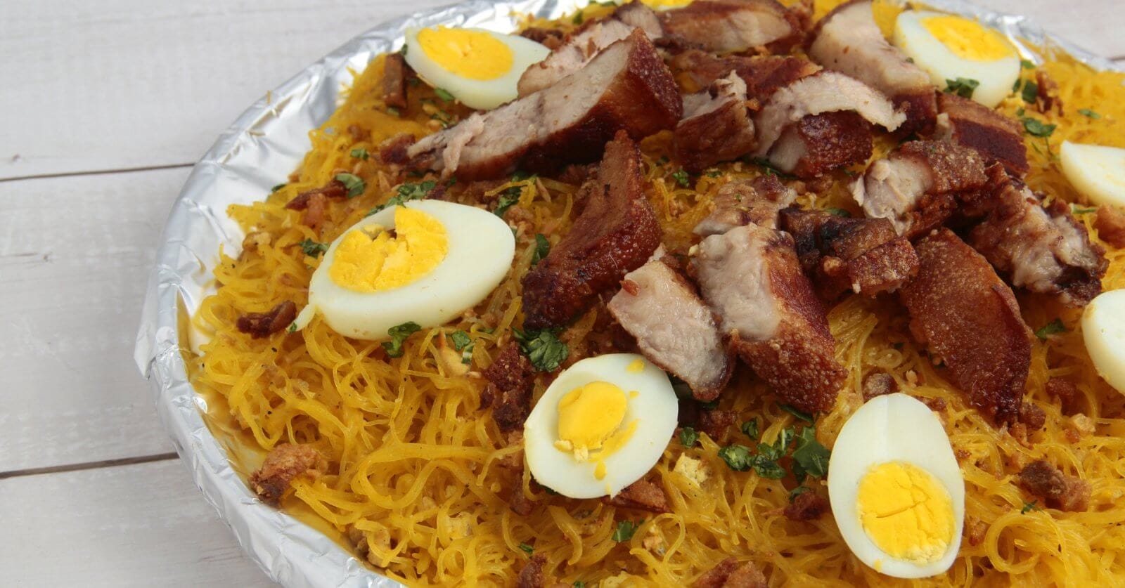 A plate filled with a Filipino noodle dish called Pancit Malabon, featuring yellow rice noodles topped with sliced hard-boiled eggs, crispy pork belly, cooked shrimp, and chopped green onions. The elements are arranged aesthetically on a white wooden table near the popular restos along Taft Avenue.