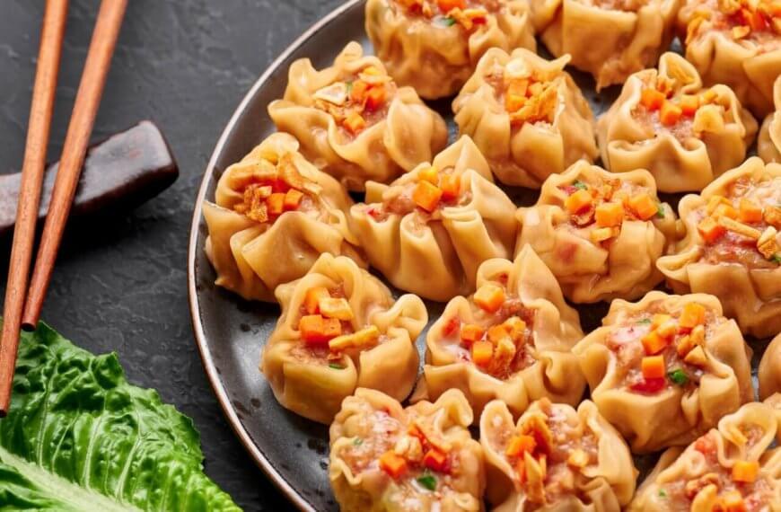 A plate filled with neatly arranged steamed dumplings, each topped with a small garnish of finely chopped vegetables. Wooden chopsticks rest on a holder beside the plate. A fresh green lettuce leaf is placed adjacent, providing a touch of color to the dark background at New Greenhills Mall's 84 restaurants.