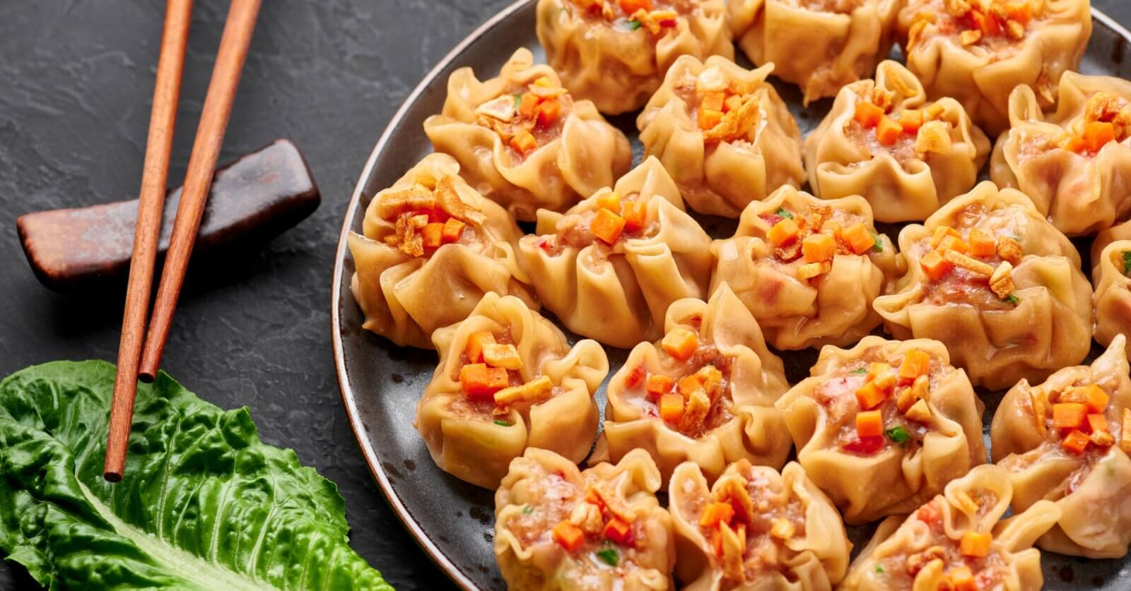 A plate filled with neatly arranged steamed dumplings, each topped with a small garnish of finely chopped vegetables. Wooden chopsticks rest on a holder beside the plate. A fresh green lettuce leaf is placed adjacent, providing a touch of color to the dark background at New Greenhills Mall's 84 restaurants.