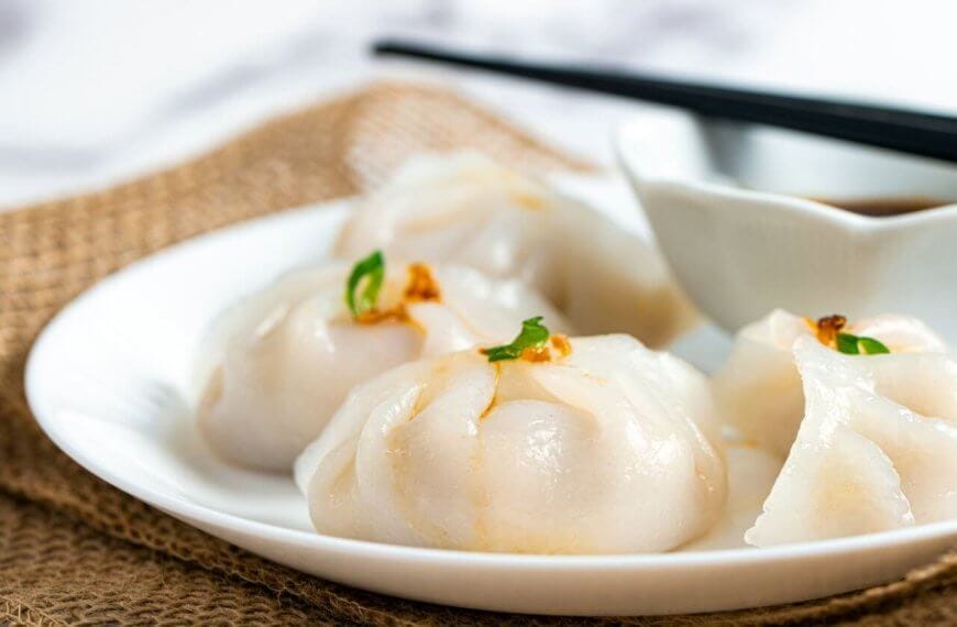 Hakaw steamed dumplings garnished with small green herbs, accompanies by a dipping sauce in a white bowl, and a pair of black chopsticks resting across it. The setup is styled on burlap fabric atop a marble surface—an elegant touch reminiscent of Top Restaurants found in Power Plant Mall.