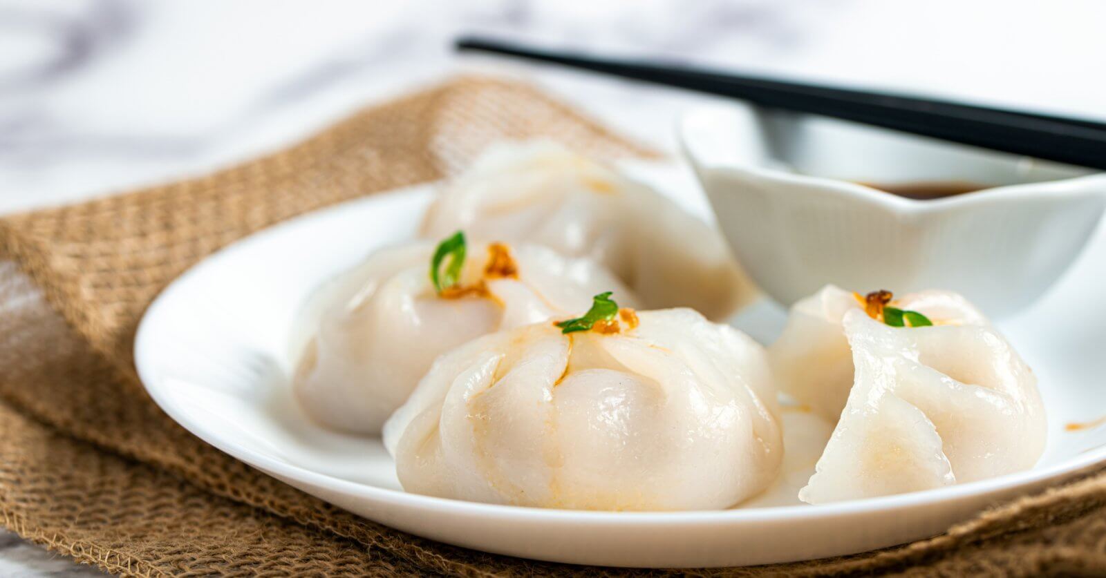 Hakaw steamed dumplings garnished with small green herbs, accompanies by a dipping sauce in a white bowl, and a pair of black chopsticks resting across it. The setup is styled on burlap fabric atop a marble surface—an elegant touch reminiscent of Top Restaurants found in Power Plant Mall.