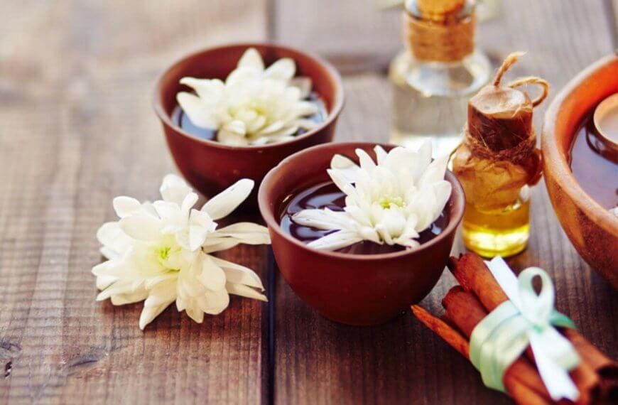 A serene spa scene on a wooden surface features two brown bowls filled with water and white daisies, a lit candle in another bowl, and a small glass bottle with a cork stopper containing oil. A bundle of cinnamon sticks tied with a green ribbon lies nearby, adding to the tranquil setup sourced from the finest fragrance suppliers in Metro Manila.