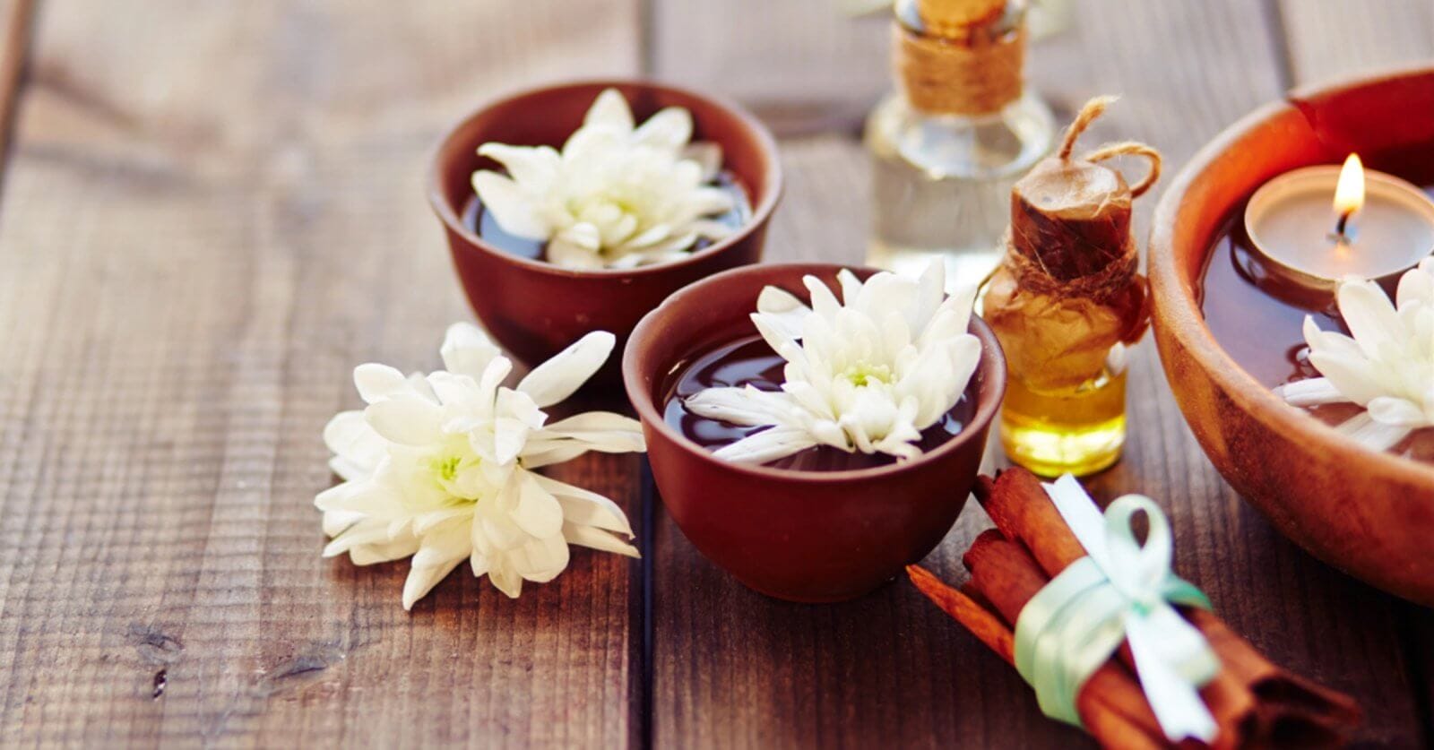 A serene spa scene on a wooden surface features two brown bowls filled with water and white daisies, a lit candle in another bowl, and a small glass bottle with a cork stopper containing oil. A bundle of cinnamon sticks tied with a green ribbon lies nearby, adding to the tranquil setup sourced from the finest fragrance suppliers in Metro Manila.