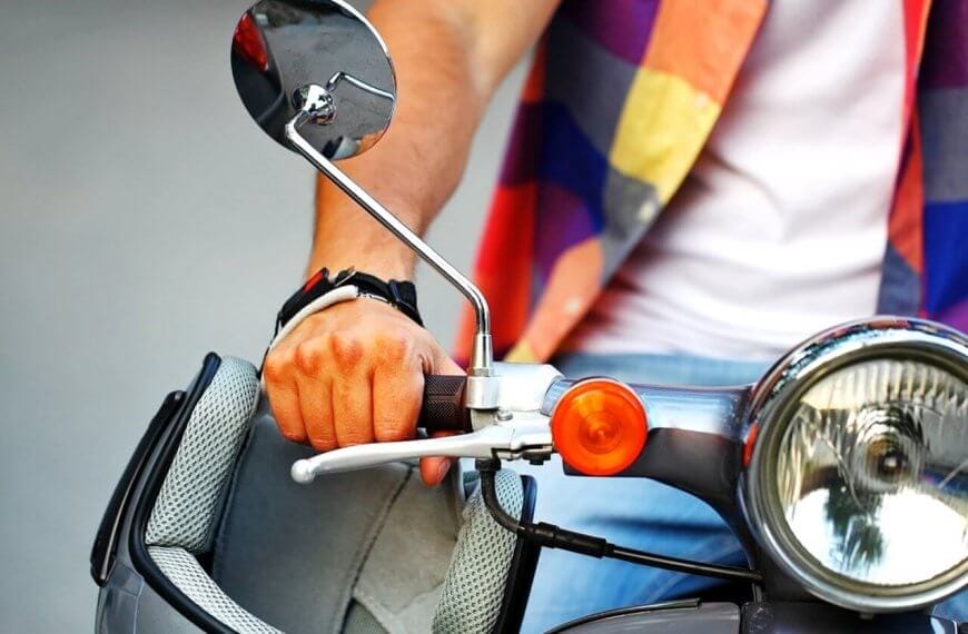 Close-up of a person wearing a colorful plaid shirt and a white T-shirt holding the handlebar of a scooter from one of the top scooter shops in Metro Manila. The focus is on their left hand gripping the handle while wearing a black wristwatch. The scooter features a large headlight, an orange turn signal, and a rearview mirror.