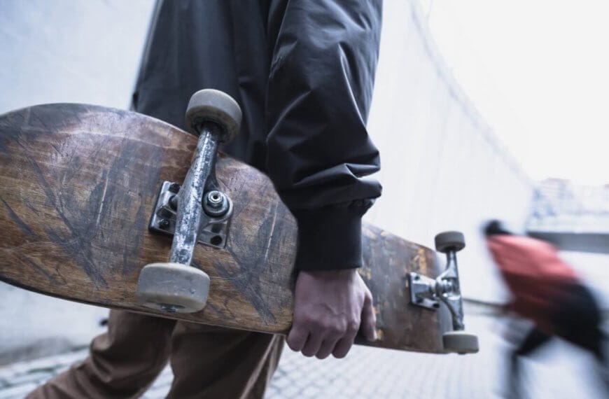 A person in a dark jacket holds a worn skateboard by their side while walking on a paved pathway. The background is a blurred image of another person in motion, wearing a red jacket, moving down a sloped area. The urban setting with concrete structures could be reminiscent of Metro Manila's bustling vibe.