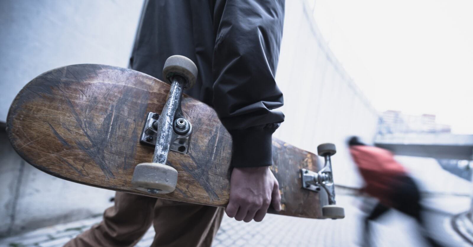 A person in a dark jacket holds a worn skateboard by their side while walking on a paved pathway. The background is a blurred image of another person in motion, wearing a red jacket, moving down a sloped area. The urban setting with concrete structures could be reminiscent of Metro Manila's bustling vibe.