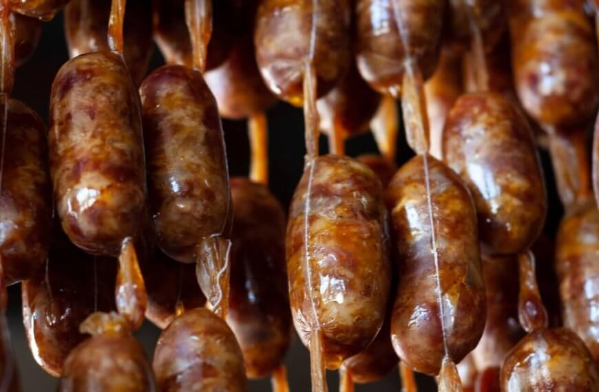 Close-up of several rows of sausages hanging vertically by beige strings. The sausages have a brownish color with visible spices embedded in the meat. The links appear glossy and slightly uneven, indicating they are likely homemade or artisanal. This delicacy could be a highlight at one of Davao's 42 Best Restaurants in SM Lanang.