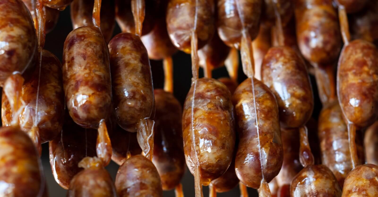 Close-up of several rows of sausages hanging vertically by beige strings. The sausages have a brownish color with visible spices embedded in the meat. The links appear glossy and slightly uneven, indicating they are likely homemade or artisanal. This delicacy could be a highlight at one of Davao's 42 Best Restaurants in SM Lanang.