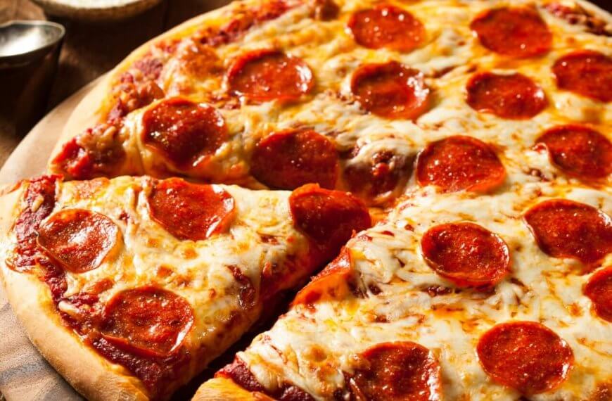 A close-up of a pepperoni pizza on a wooden surface. The pizza has a golden-brown crust and is topped with melted cheese and evenly distributed pepperoni slices. One slice is partially separated from the rest, showcasing the gooey cheese and crisp edges. Leafy greens and utensils are blurred in the background, reminiscent of a Sulit Dining Experience at SM Manila's best restaurants.